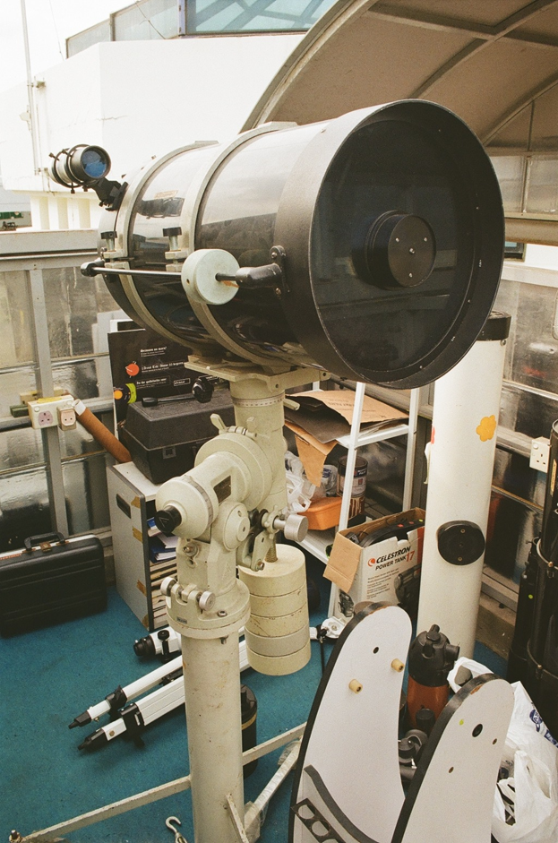 A large motorized telescope mounted on a tracking stand inside an observatory or equipment room, with cables and mechanical components visible.
